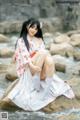 A woman in a kimono sitting on a rock by the water.